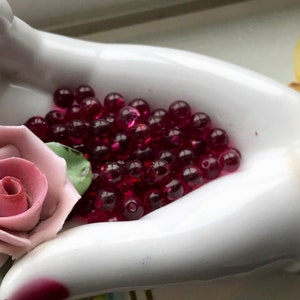May include: A white porcelain hand holding a large quantity of deep red glass beads. The hand is decorated with a pink rose.