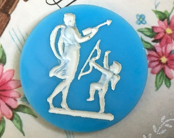 May include: A blue glass button with a white cameo design of a woman and a cherub. The woman is holding a bow and arrow, and the cherub is holding a bow and arrow.
