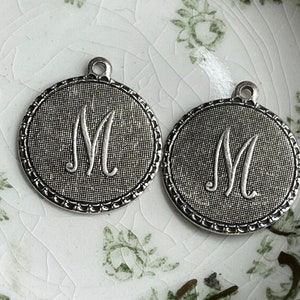 May include: Two silver-toned metal charms with a beaded edge, each featuring a raised letter "M" in a circular design.