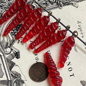 May include: A string of seven red glass beads, each with a spiral design. The beads are strung on a wire with a brass loop at the end of each bead. The beads are lying on a book with a black and white illustration.