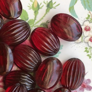 May include: A collection of 25 oval-shaped, red glass beads with a ribbed texture. The beads are arranged in a pile on a floral patterned surface.