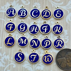 May include: A set of 26 gold-colored metal charms with blue enamel and white letters. Each charm features a different letter of the alphabet, from A to Z. The charms are approximately the size of a US penny.