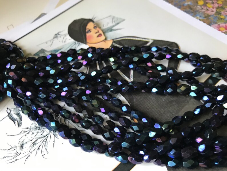 May include: A strand of dark blue iridescent glass beads. The beads are faceted and have a slightly oval shape.