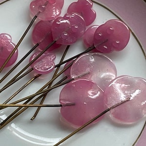 May include: A collection of pink glass flower head pins with gold metal stems. The flower heads are translucent and have a slightly iridescent finish.