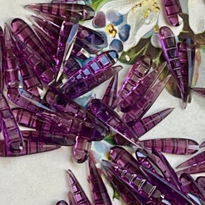 May include: A large group of purple glass teardrop-shaped beads with a textured surface. The beads are arranged in a random pattern on a white background.