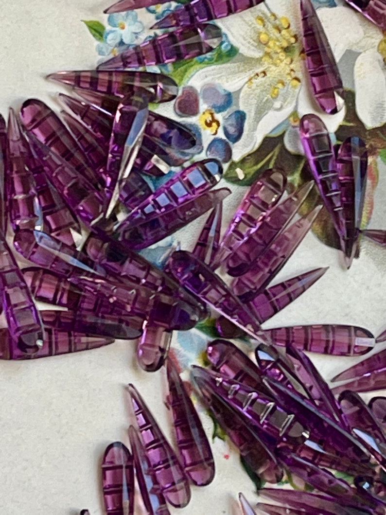 May include: A large group of purple glass teardrop-shaped beads with a textured surface. The beads are arranged in a random pattern on a white background.