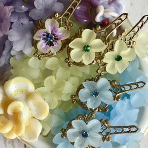 May include: Assortment of glass flower charms with gold metal accents. The charms feature a variety of colors, including purple, yellow, blue, and green. The charms are arranged in a white bowl.