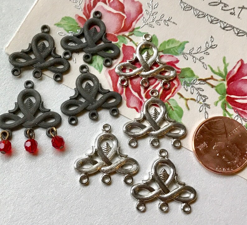 May include: A collection of silver and black metal charms with intricate designs. The charms have three loops at the bottom and a decorative top. Some of the charms have red glass beads attached.