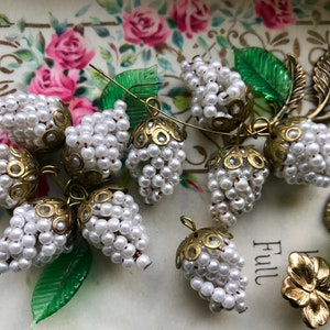 May include: A cluster of eight white pearl and gold bead clusters, each with a delicate gold filigree design. The clusters are arranged on a white surface with green leaves and a gold floral embellishment.