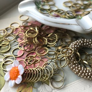 Peut inclure: Gros plan sur un tas d'anneaux de saut dor&eacute;s, un plat en porcelaine blanche avec des bordures dor&eacute;es et un bracelet en perles beiges. Une petite fleur blanche avec un centre orange est au premier plan.