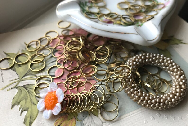Peut inclure: Gros plan sur un tas d'anneaux de saut dor&eacute;s, un plat en porcelaine blanche avec des bordures dor&eacute;es et un bracelet en perles beiges. Une petite fleur blanche avec un centre orange est au premier plan.