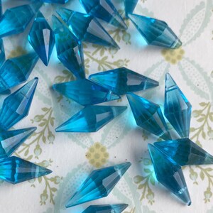 May include: A scattering of turquoise blue faceted glass teardrop beads. The beads are arranged on a white background with a floral pattern.