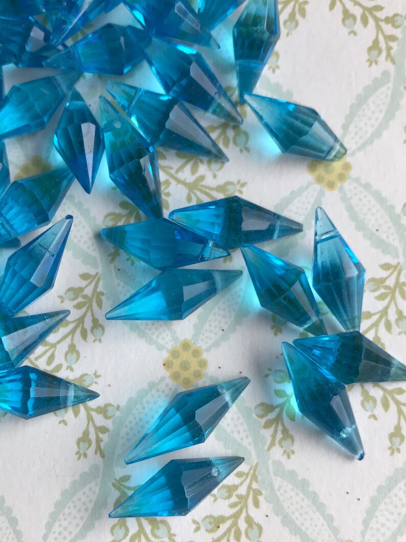 May include: A scattering of turquoise blue faceted glass teardrop beads. The beads are arranged on a white background with a floral pattern.