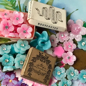 May include: Assortment of vintage pink and blue glass buttons with rhinestones. The buttons are in small boxes with French text that reads "Grand Nouveautés".