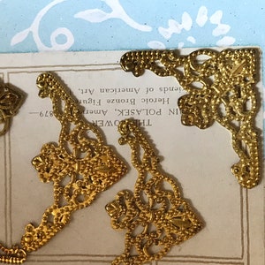 May include: Gold metal corner embellishments with a delicate, intricate design. The embellishments have a beaded edge and are perfect for adding a touch of elegance to craft projects.
