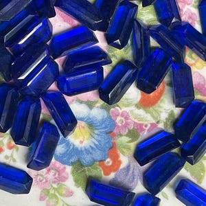 May include: A collection of numerous small, rectangular, faceted deep blue glass beads. Each bead features a polished surface with sharp edges and a uniform, intense cobalt blue color, scattered across a surface with a floral pattern.