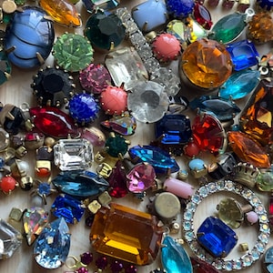 May include: A selection of vintage rhinestones and jewels in diverse shapes and sizes. The collection features round, oval, and rectangular stones in blue, green, pink, orange, and clear hues. Some are set in gold-tone metal.
