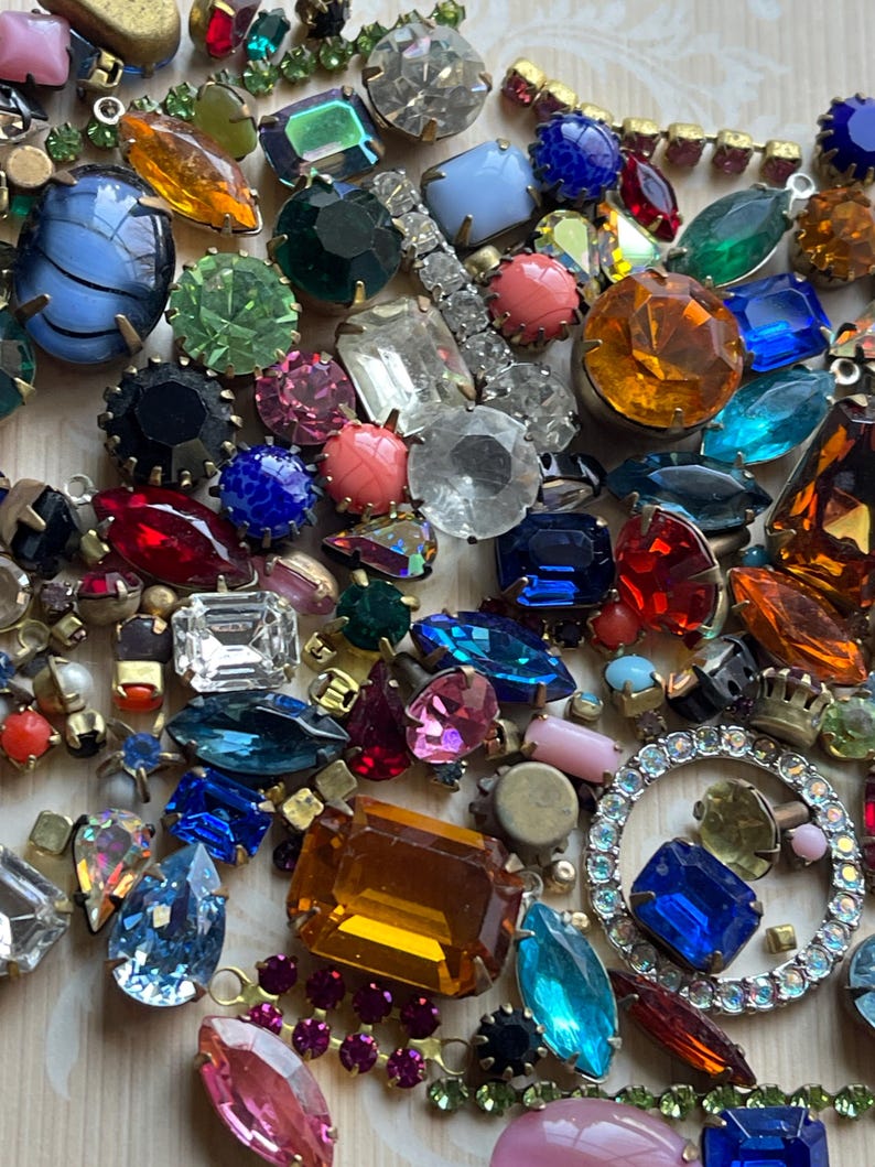 May include: A selection of vintage rhinestones and jewels in diverse shapes and sizes. The collection features round, oval, and rectangular stones in blue, green, pink, orange, and clear hues. Some are set in gold-tone metal.