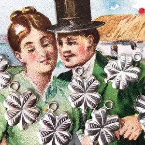 May include: Seven silver four-leaf clover charms with a loop for hanging. The charms are arranged on a vintage postcard featuring a couple in front of a thatched roof cottage.