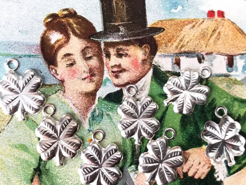 May include: Seven silver four-leaf clover charms with a loop for hanging. The charms are arranged on a vintage postcard featuring a couple in front of a thatched roof cottage.