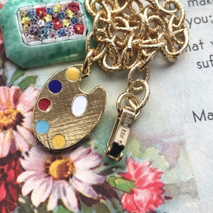 K&ouml;nnte beinhalten: Eine goldene Kettenkette mit einem Anh&auml;nger in Form eines gr&uuml;nen und wei&szlig;en Mosaik-Bilderrahmens und einem goldenen Malpalette-Anh&auml;nger mit bunten Emaille-Farbtupfern. Die Kette hat einen Verschluss mit der Aufschrift "Made in Italy".