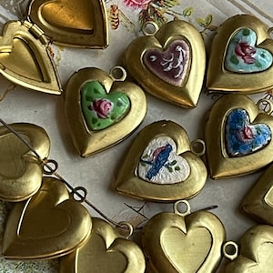 May include: A collection of 10 gold heart-shaped locket pendants with various painted designs. Some lockets feature floral designs, while others have birds or other motifs. The lockets are arranged on a patterned surface.