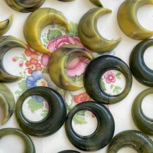 May include: An assortment of vintage, olive green and yellow-toned crescent moon and circular shaped beads. The beads have a marbled appearance and are arranged on a floral patterned surface.