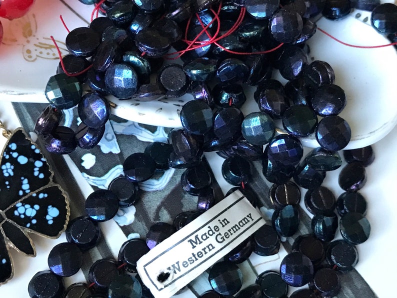 Puede incluir: Una hebra de cuentas de vidrio negro iridiscente con acabado facetado. Las cuentas est&aacute;n enhebradas en un hilo rojo y tienen una etiqueta que dice "Made in Western Germany".