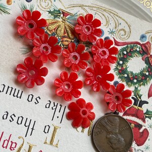 May include: Nine red plastic flower-shaped beads with a hole in the center. The beads are arranged in a semi-circle on a white surface with a gold border.