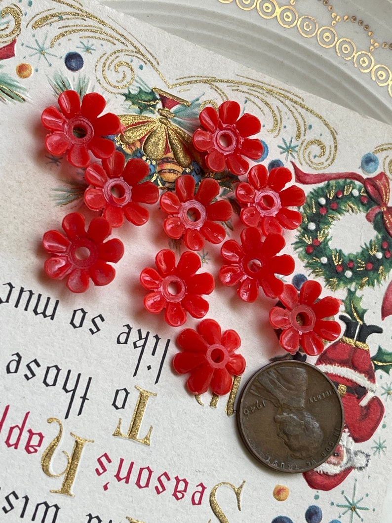 May include: Nine red plastic flower-shaped beads with a hole in the center. The beads are arranged in a semi-circle on a white surface with a gold border.