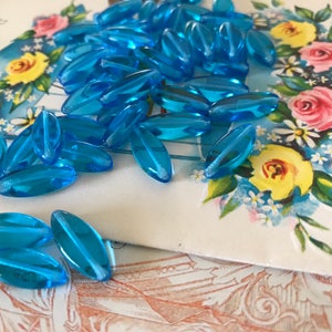 May include: A pile of light blue glass beads in an oval shape. The beads are on a white card with a floral design.