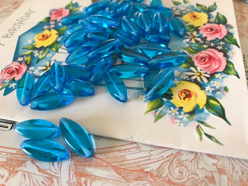 May include: A pile of light blue glass beads in an oval shape. The beads are on a white card with a floral design.
