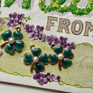 Puede incluir: Tres colgantes de tr&eacute;bol de cuatro hojas dorados con esmalte verde y una perla central. Los colgantes est&aacute;n colocados sobre una postal vintage con elementos florales y de texto. El fondo presenta flores moradas y follaje verde.