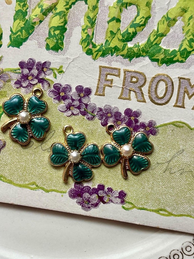 Puede incluir: Tres colgantes de tr&eacute;bol de cuatro hojas dorados con esmalte verde y una perla central. Los colgantes est&aacute;n colocados sobre una postal vintage con elementos florales y de texto. El fondo presenta flores moradas y follaje verde.