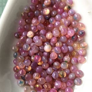 K&ouml;nnte beinhalten: Ein Haufen rosa Opal-Cabochons mit irisierendem Schimmer. Die Cabochons sind rund und glatt und in einem zuf&auml;lligen Muster angeordnet.