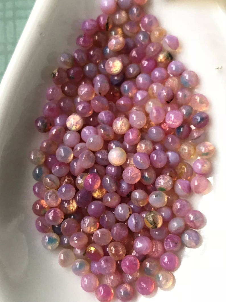 K&ouml;nnte beinhalten: Ein Haufen rosa Opal-Cabochons mit irisierendem Schimmer. Die Cabochons sind rund und glatt und in einem zuf&auml;lligen Muster angeordnet.