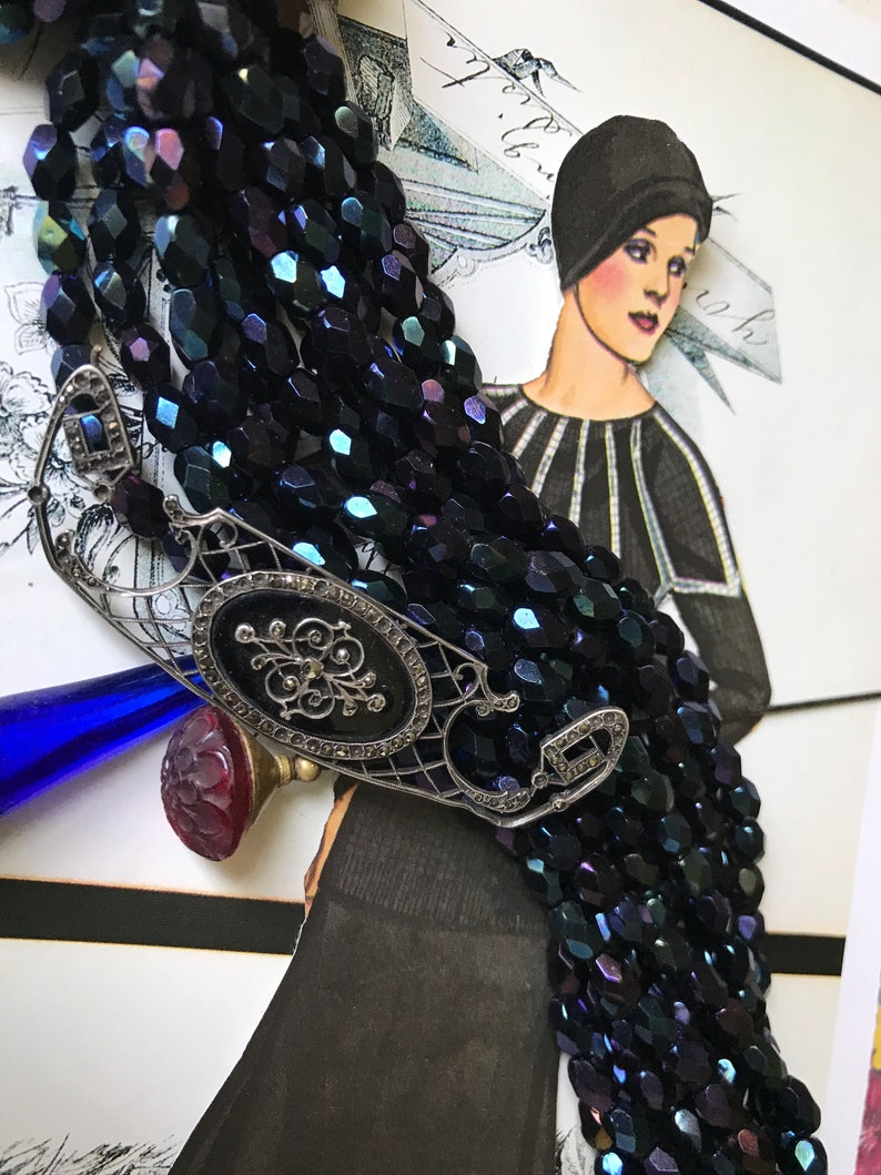 May include: A multi-strand necklace made of dark blue faceted glass beads with a silver filigree pendant featuring a black enamel oval and rhinestones. The pendant is attached to a red cameo.