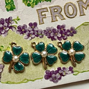 Puede incluir: Tres colgantes de tr&eacute;bol de cuatro hojas de esmalte verde con detalles dorados y una perla central. Los colgantes est&aacute;n colocados sobre una postal vintage con elementos florales y de texto. Los colgantes de tr&eacute;bol son un s&iacute;mbolo de buena suerte.