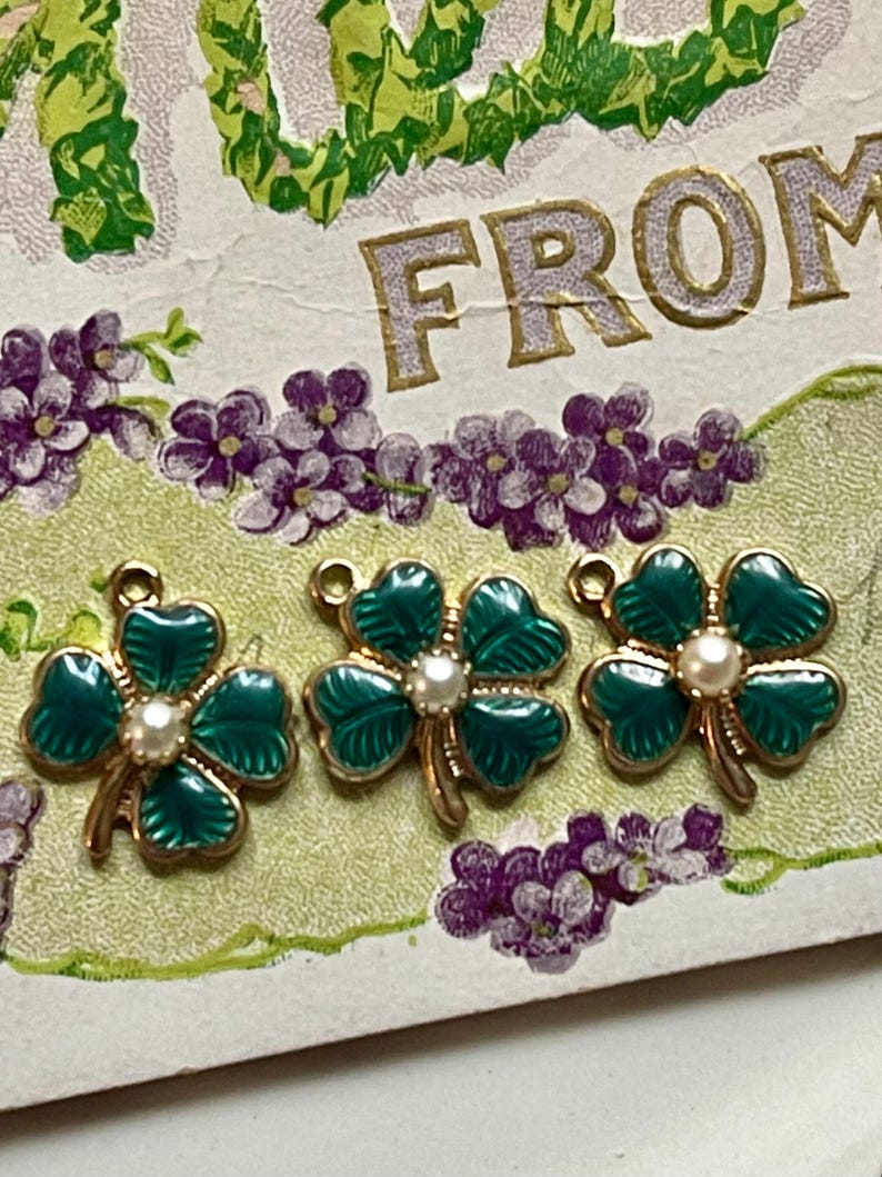 Puede incluir: Tres colgantes de tr&eacute;bol de cuatro hojas de esmalte verde con detalles dorados y una perla central. Los colgantes est&aacute;n colocados sobre una postal vintage con elementos florales y de texto. Los colgantes de tr&eacute;bol son un s&iacute;mbolo de buena suerte.