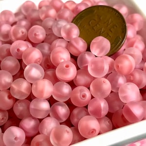 May include: A close-up of a pile of pink glass beads. The beads are round and have a frosted finish. A US penny is included for scale.