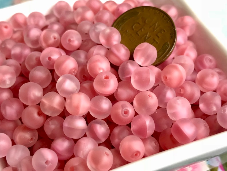 May include: A close-up of a pile of pink glass beads. The beads are round and have a frosted finish. A US penny is included for scale.