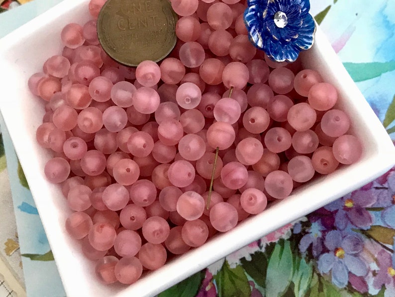 May include: A bowl filled with translucent pink beads. The beads are round and have a frosted finish.