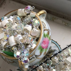 Puede incluir: Un jarr&oacute;n de porcelana blanca con ribetes dorados y un dise&ntilde;o de rosa rosa, lleno de una cadena de cuentas de cristal iridiscente con etiquetas de papel adjuntas. Las etiquetas tienen palabras escritas a mano, incluyendo "Jan", "Nov" y "Dec".