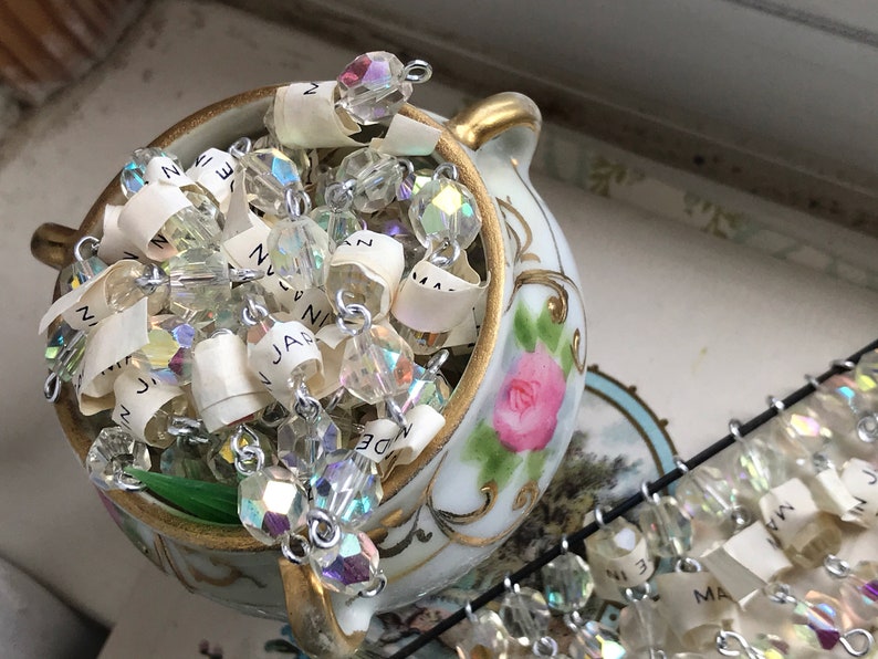 Puede incluir: Un jarr&oacute;n de porcelana blanca con ribetes dorados y un dise&ntilde;o de rosa rosa, lleno de una cadena de cuentas de cristal iridiscente con etiquetas de papel adjuntas. Las etiquetas tienen palabras escritas a mano, incluyendo "Jan", "Nov" y "Dec".