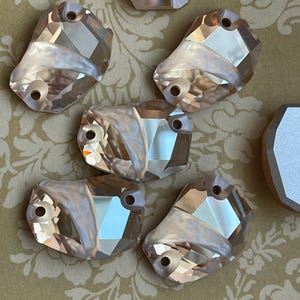 May include: Close-up of several faceted, light peach-colored crystal embellishments. Each has two holes for attachment and a pearlescent, curved accent. The crystals are arranged on a floral patterned background.