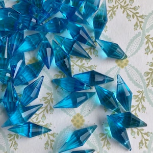 May include: A pile of turquoise blue faceted glass beads in a diamond shape. The beads are scattered on a white background with a floral pattern.