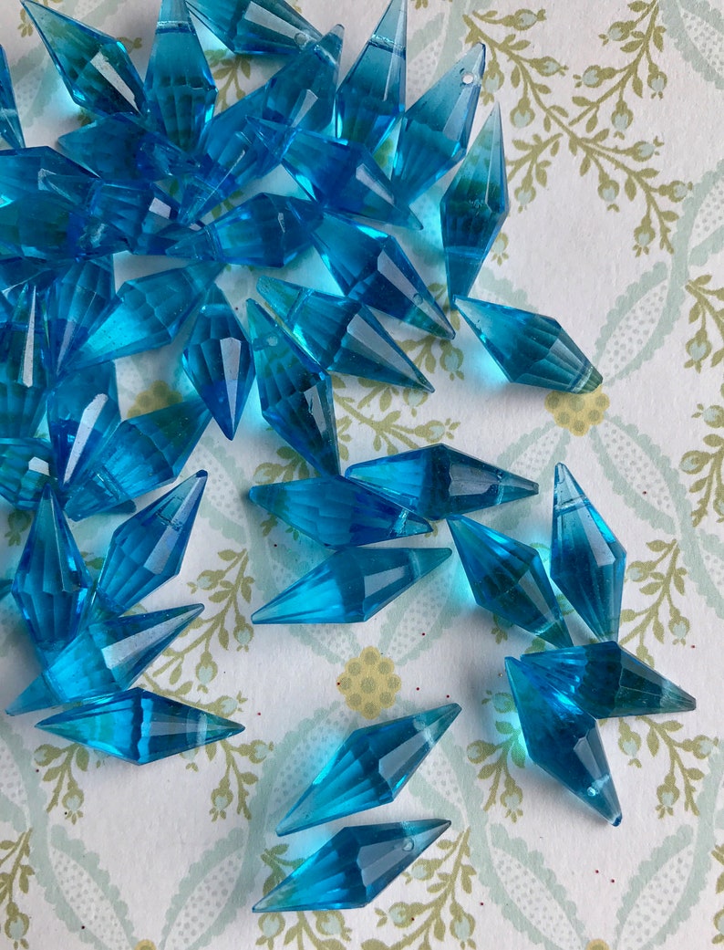 May include: A pile of turquoise blue faceted glass beads in a diamond shape. The beads are scattered on a white background with a floral pattern.