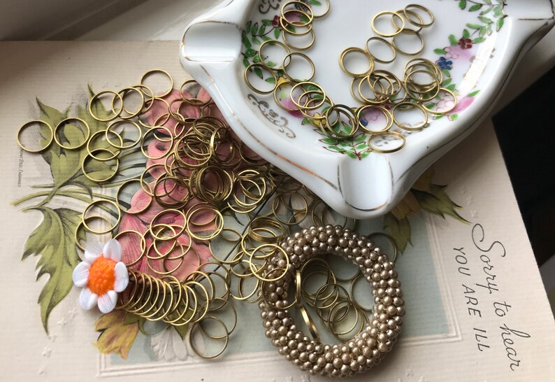 Peut inclure: Un tas d'anneaux de saut dor&eacute;s, certains dans un plat en porcelaine blanche avec des motifs floraux, et une couronne de perles dor&eacute;es sur une carte vintage qui dit "D&eacute;sol&eacute; d'apprendre que vous &ecirc;tes malade".