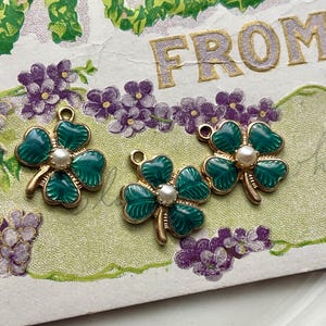 Puede incluir: Tres dijes de tr&eacute;bol de cuatro hojas verdes y dorados con un centro de perla se muestran en una postal vintage con flores moradas. Los dijes tienen un tallo dorado y un lazo para sujetar a joyas o manualidades.