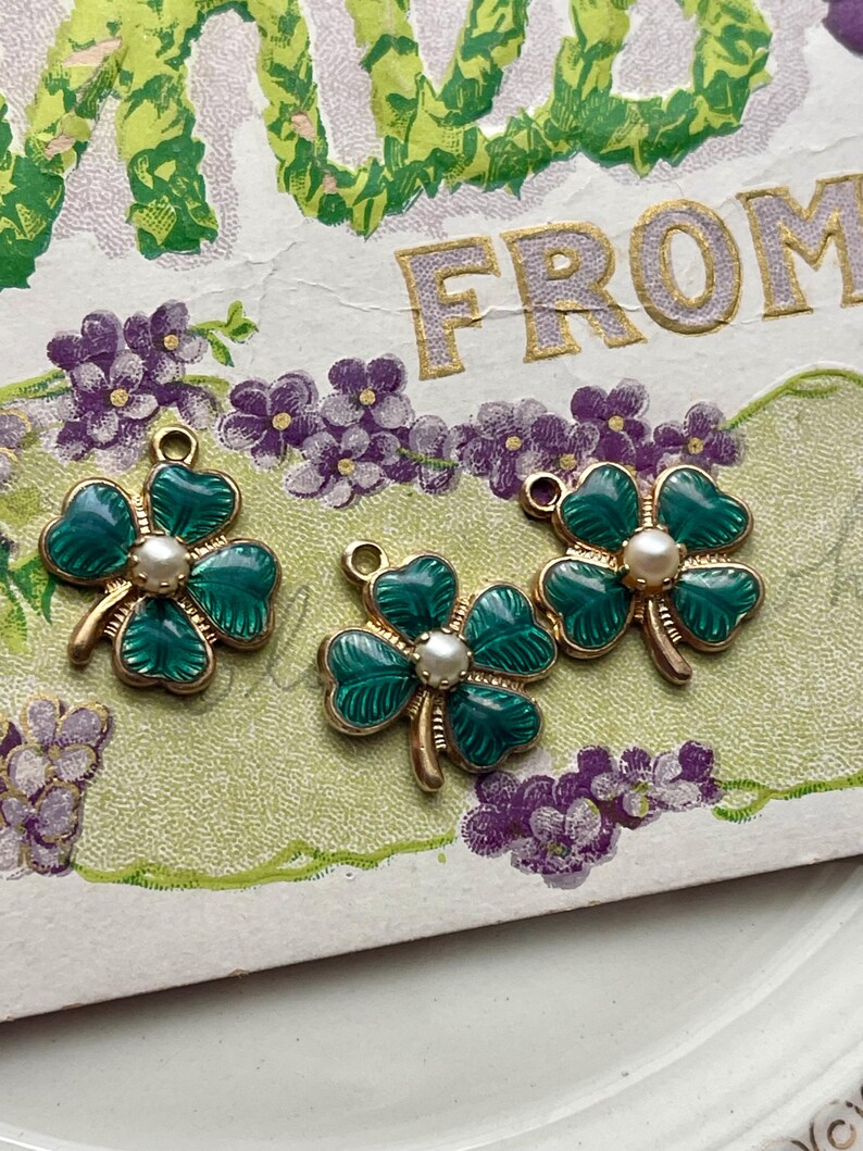 Puede incluir: Tres dijes de tr&eacute;bol de cuatro hojas verdes y dorados con un centro de perla se muestran en una postal vintage con flores moradas. Los dijes tienen un tallo dorado y un lazo para sujetar a joyas o manualidades.